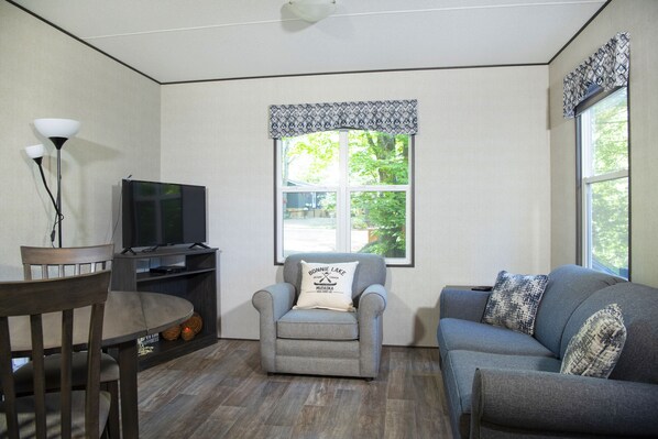 Una sala de estar con un sofá gris, un sillón gris, un televisor sobre un mueble bajo, una mesa de comedor con sillas y un ventanal con cortinas estampadas.