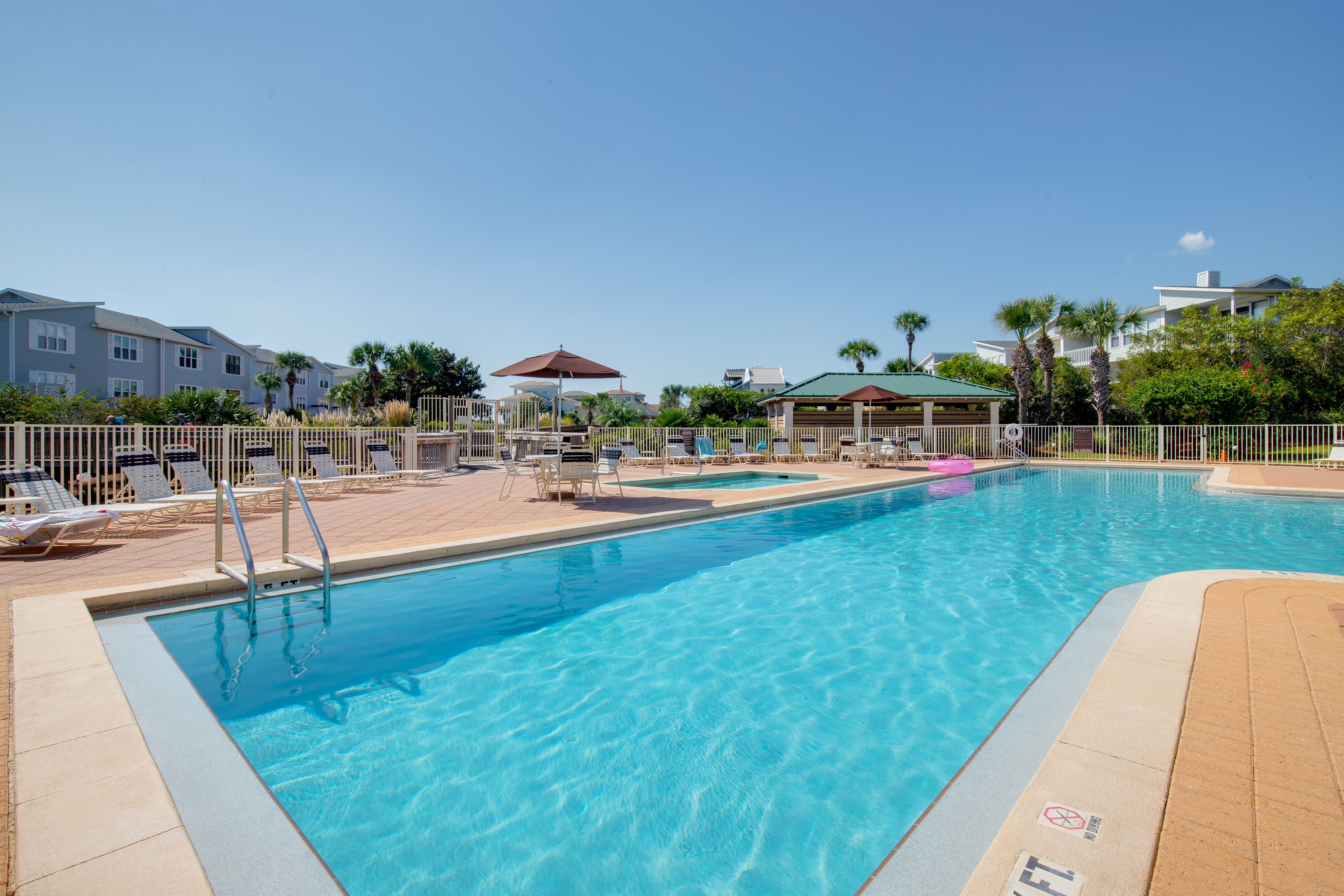 A swimming pool with lounge chairs and umbrellas, surrounded by a fence and palm trees.