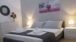 Een net opgemaakt bed met wit en grijs beddengoed, twee nachtkastjes met lampen, een wandspiegel en een schilderij van een boom.