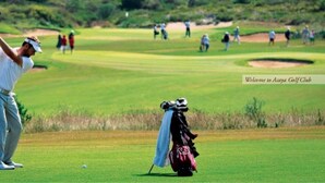 Een golfbaan met een speler in een witte blouse en grijze broek, die zich klaarmaakt om een golfbal te slaan.