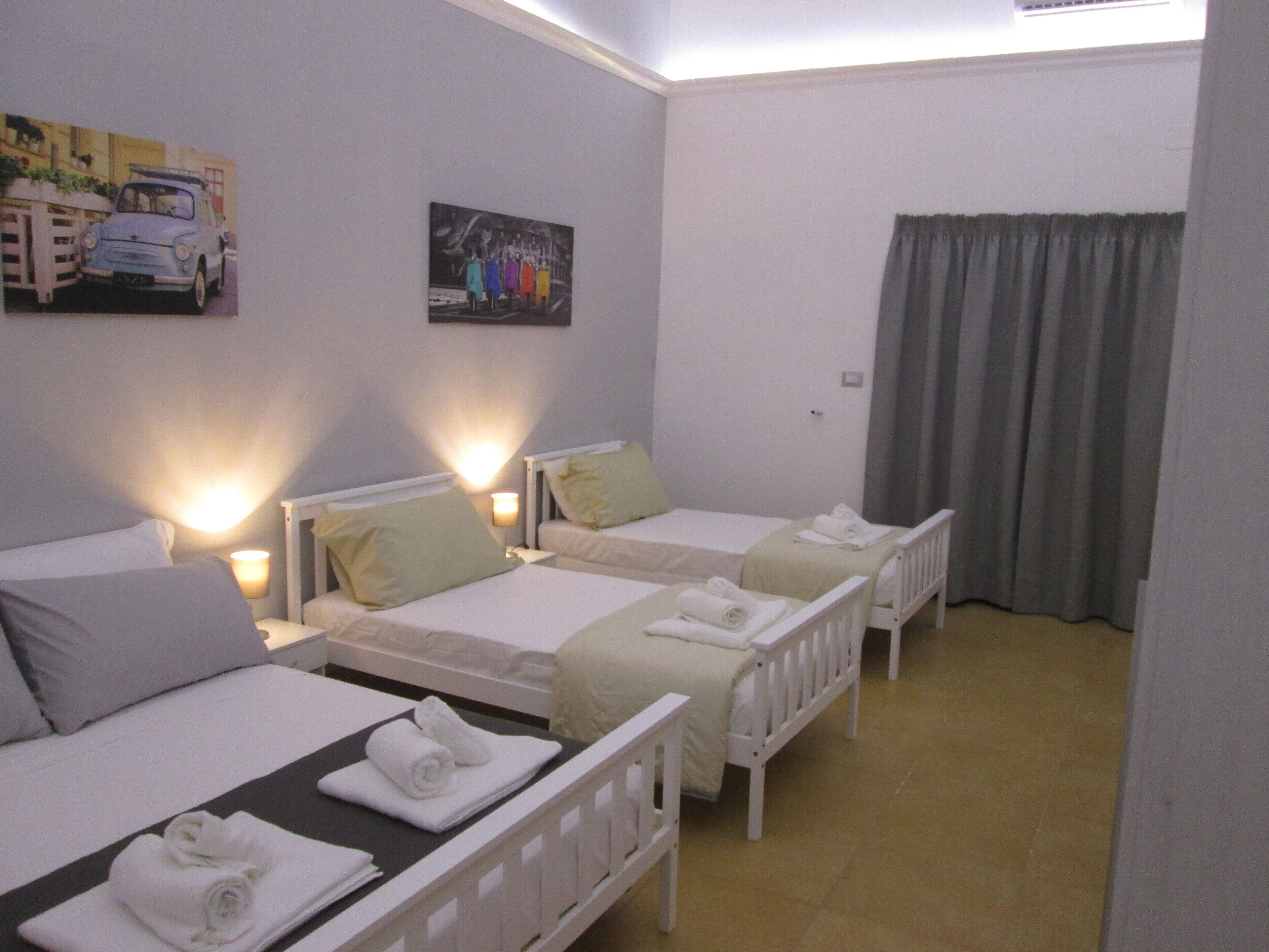 Une chambre d’hôtel avec trois lits simples, chacun doté d’une tête de lit blanche et d’un oreiller. Un tableau représentant une voiture est accroché au mur au-dessus du lit situé à gauche, et un tableau représentant un groupe de personnes est accroché au mur au-dessus du lit situé au centre. La chambre est équipée d’une fenêtre avec des rideaux, d  ’un radiateur et d’une petite table avec une lampe.