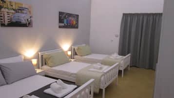 Ein Hotelzimmer mit drei Einzelbetten, jeweils mit weißem Kopfteil und Kissen. Über dem linken Bett hängt ein Bild von einem Auto, über dem mittleren Bett ein Gemälde einer Personengruppe. Das Zimmer verfügt über ein Fenster mit Vorhängen, eine Heizung und einen kleinen Tisch mit einer Lampe.