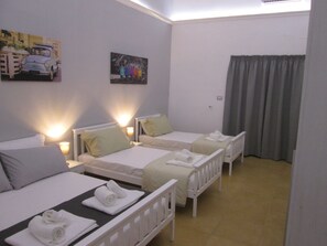 A hotel room with three single beds, each with a white headboard and a pillow. There is a painting of a car on the wall above the left bed, and a painting of a group of people on the wall above the middle bed. The room has a window with curtains, a radiator, and a small table with a lamp.