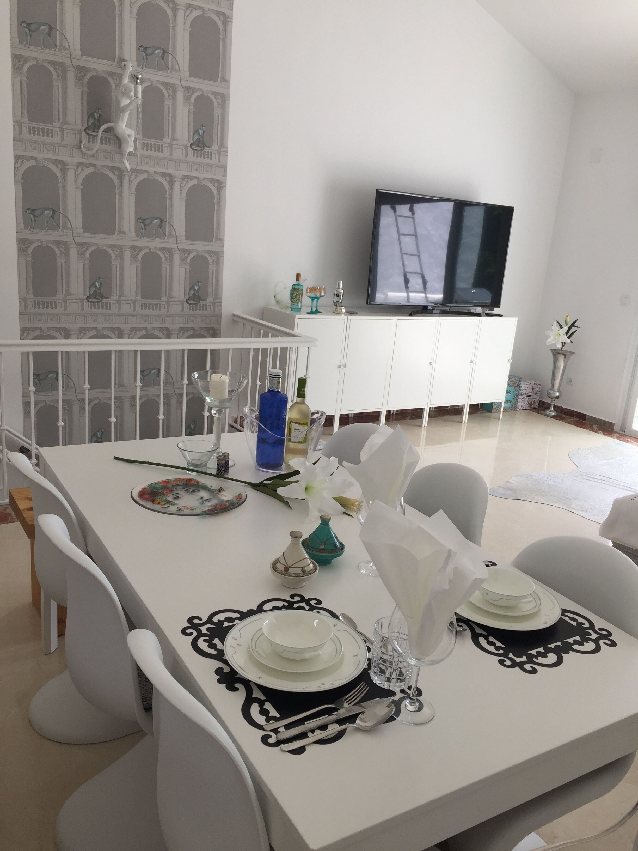 A dining area with a white table set for two, a TV on a white cabinet, and a balcony with a view of a building with arches.