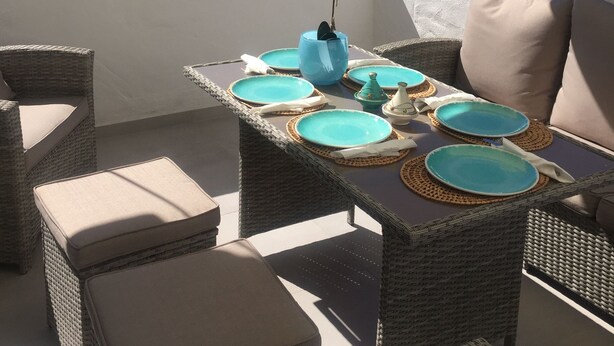 Una terrazza con mobili in vimini, un tavolo apparecchiato con piatti blu e vista sul mare e sul paesaggio.