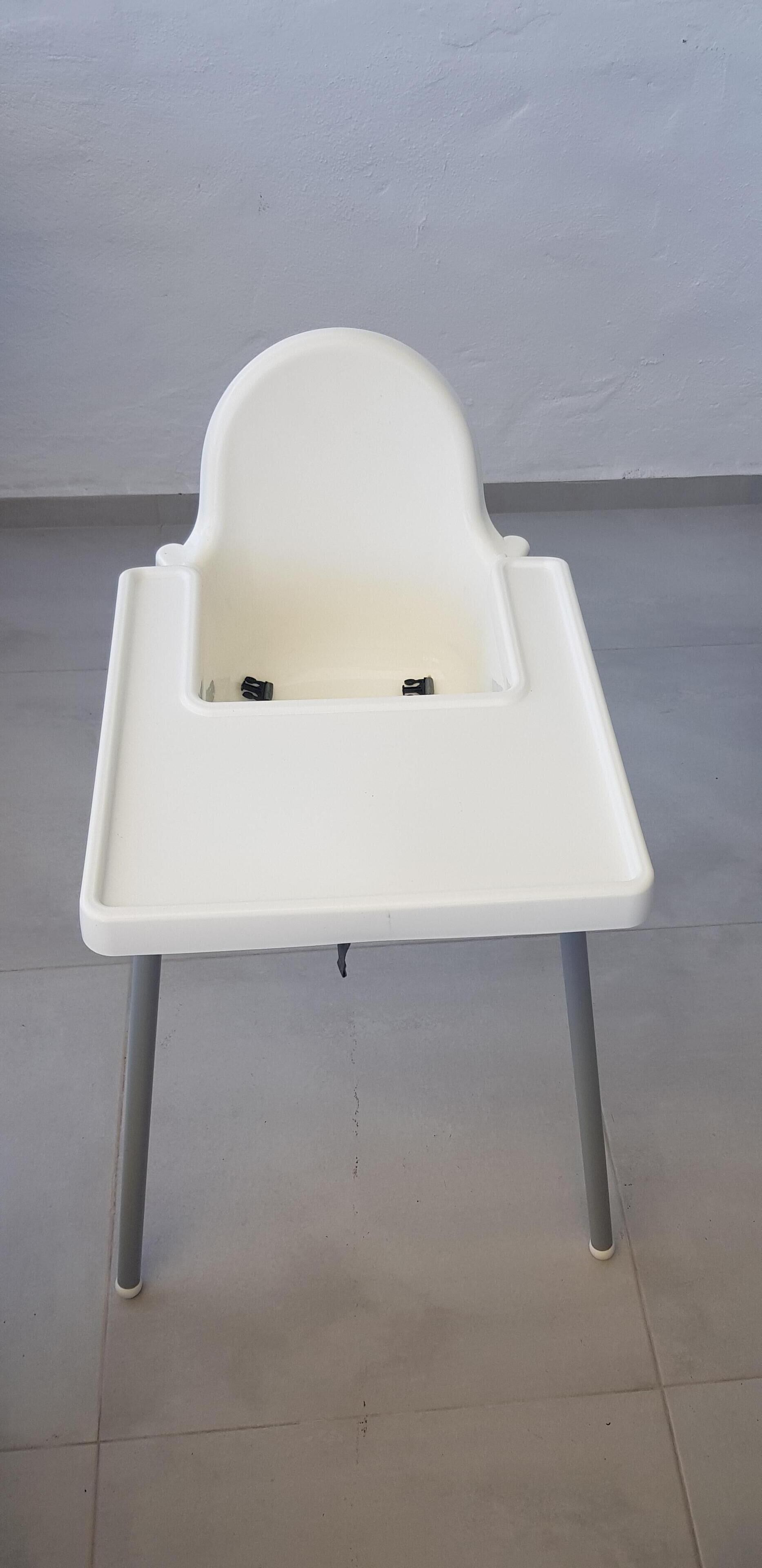A white high chair with a tray table, positioned on a tiled floor against a plain wall.