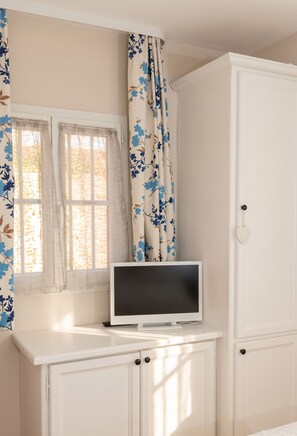 A room with a television on a white cabinet, a window with floral curtains, and a white wardrobe. - Homerez - Apt near beach & pool (Six-Fours-les-Plages)