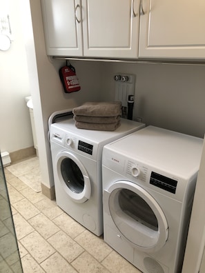 A laundry area with two stacked front-loading washing machines and a dryer, a white tiled floor, and a red fire extinguisher mounted on the wall.