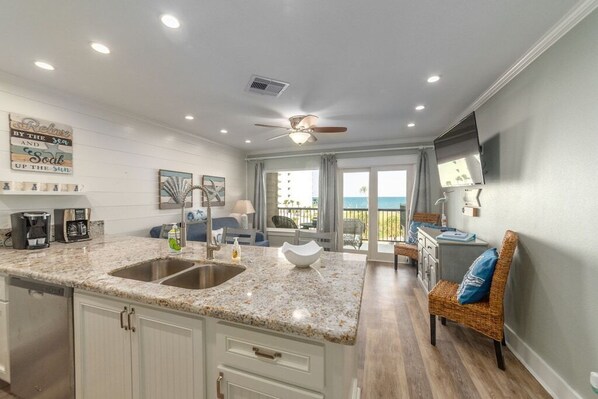 A kitchen with granite countertops, white cabinetry, and a large island with a sink. There is a dining area with a wicker chair and a view of the ocean.