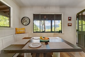 A dining area with a large window, a clock, and a bowl of fruit on the table. - 3BR cabin with hot tub, mtn views, nearby lake access; near Tail of the Dragon; free rafting & train (Robbinsville)