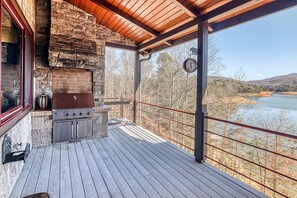 A wooden deck with a stone fireplace, a clock, and a view of a lake and hills. - Fontana Lake Lodge: 4BR/4BA lakefront home with hot tub, private dock, game room (Bryson City)