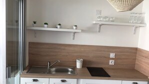 A modern kitchen with white cabinets, a stainless steel sink, and a wooden countertop. There are two white shelves above the countertop, each holding small potted plants and glassware. A woven pendant light hangs above the countertop.
