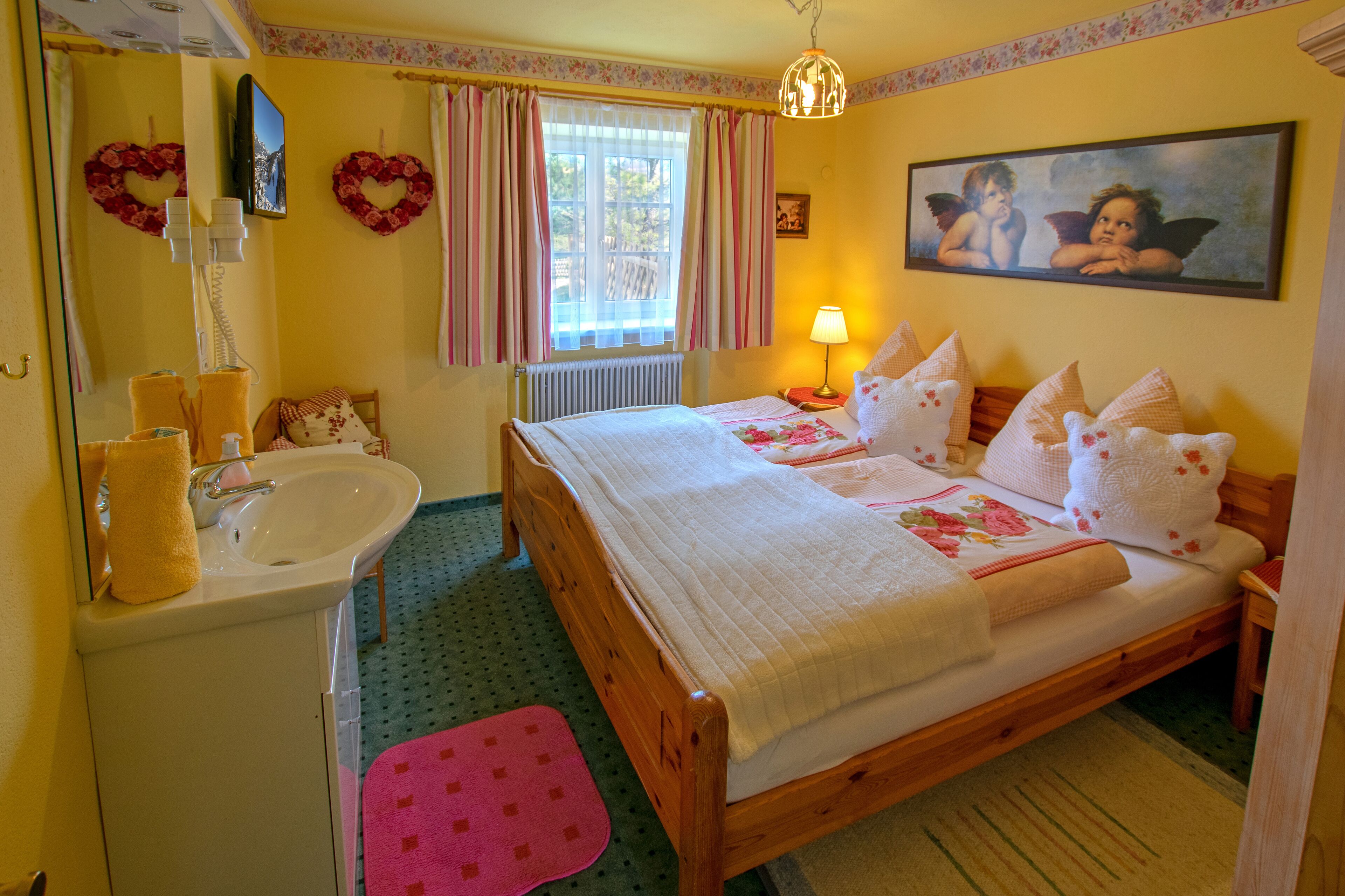 A bedroom with a wooden bed, a bedside table, a lamp, a mirror, a sink, and a window with curtains.