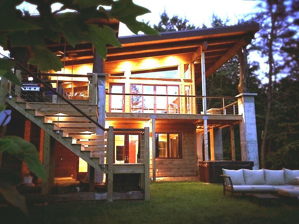 A two-story wooden cabin with a staircase leading to the upper level, surrounded by a grassy yard and trees.