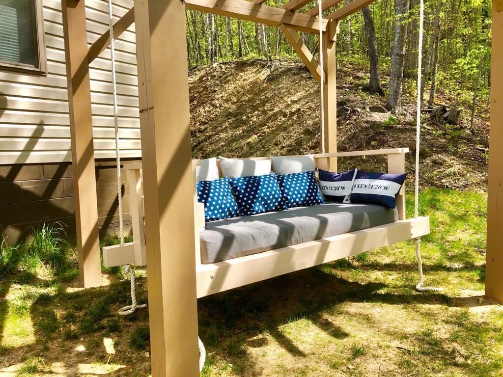Un lit-balançoire en bois, agrémenté d’un coussin et de plusieurs oreillers, installé devant un décor de forêt.