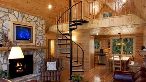 A wooden cabin interior with a stone fireplace, a spiral staircase, a dining area, and a kitchen.