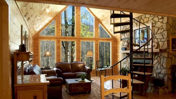 A cozy cabin interior with a wooden interior, a stone fireplace, a leather sofa, and a spiral staircase.