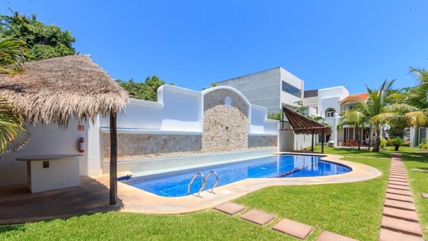 Una piscina con una pared de piedra y una estructura con techo de paja, rodeada de un césped cuidado y un camino pavimentado.
