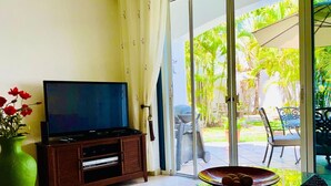 Una sala de estar con un televisor sobre un mueble de madera, una mesa de centro de vidrio con un frutero y un jarrón verde con flores rojas.