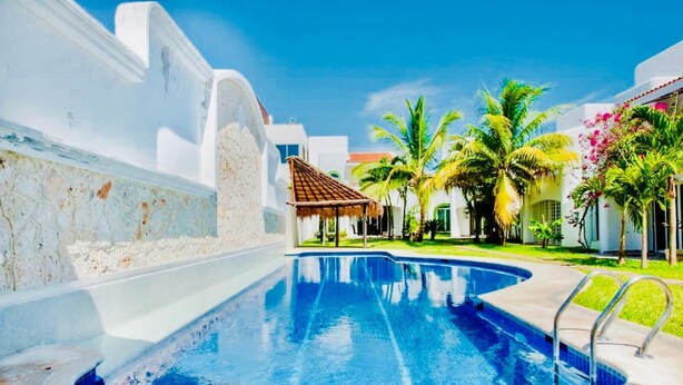 A swimming pool with a thatched-roof structure, surrounded by palm trees and white buildings. - Dolce Vita Caribe Executive Triple Room with Pool – Self Check-in - Playacar (Playa del Carmen)