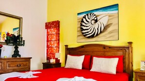 A hotel room with a red bedspread, white pillows, a wooden headboard, a wall-mounted zebra shell artwork, a mirror, a red floral curtain, and a wooden nightstand with a lamp. - Dolce Vita Caribe Executive Triple Room with Pool – Self Check-in - Playacar (Playa del Carmen)