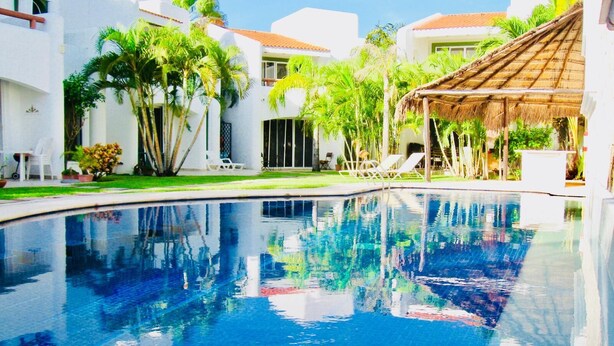 A swimming pool with clear blue water, surrounded by white buildings with red-tiled roofs, palm trees, and lounge chairs.