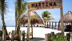 A wooden sign with the text "MARTINA BEACH" and thatch-roofed structures, palm trees, and beach umbrellas.