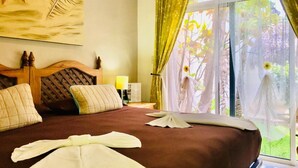 A hotel room with a large bed, a wooden headboard, a nightstand with a lamp, and a view of greenery through the window.