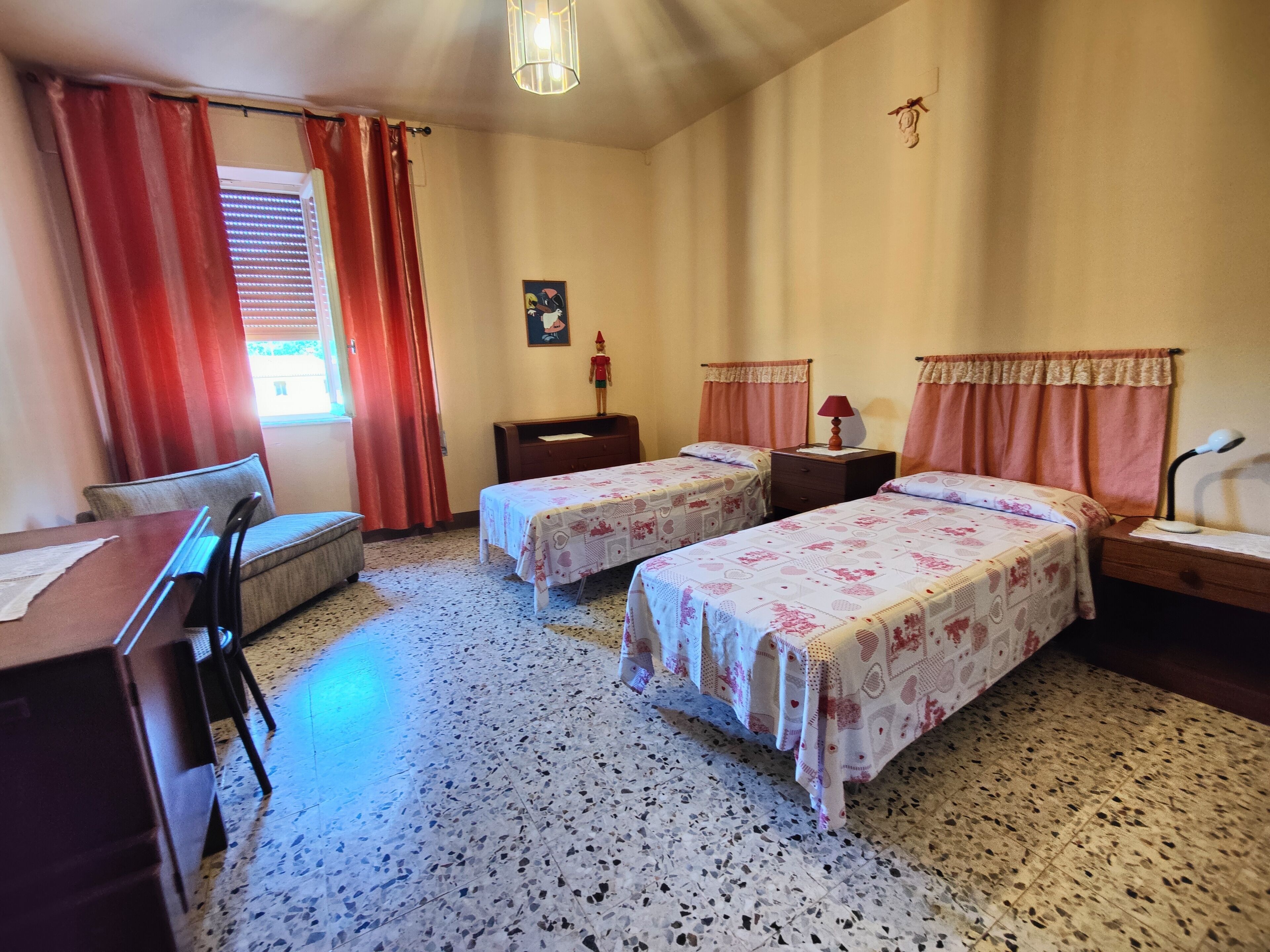 Una stanza con due letti, una scrivania, una sedia, un tavolino, una lampada e una finestra con tende.