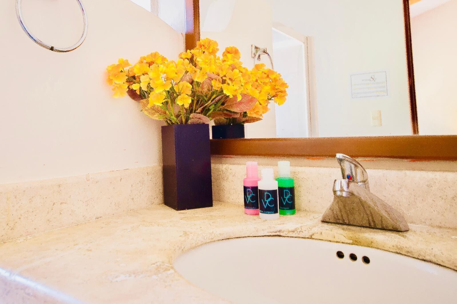 Een badkamer met een wastafel, spiegel en een vaas met gele bloemen. Drie flessen staan naast de spiegel.