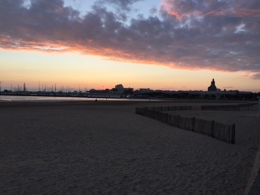 Una spiaggia con una recinzione di legno e sullo sfondo, al tramonto, la linea dell'orizzonte di una città.
