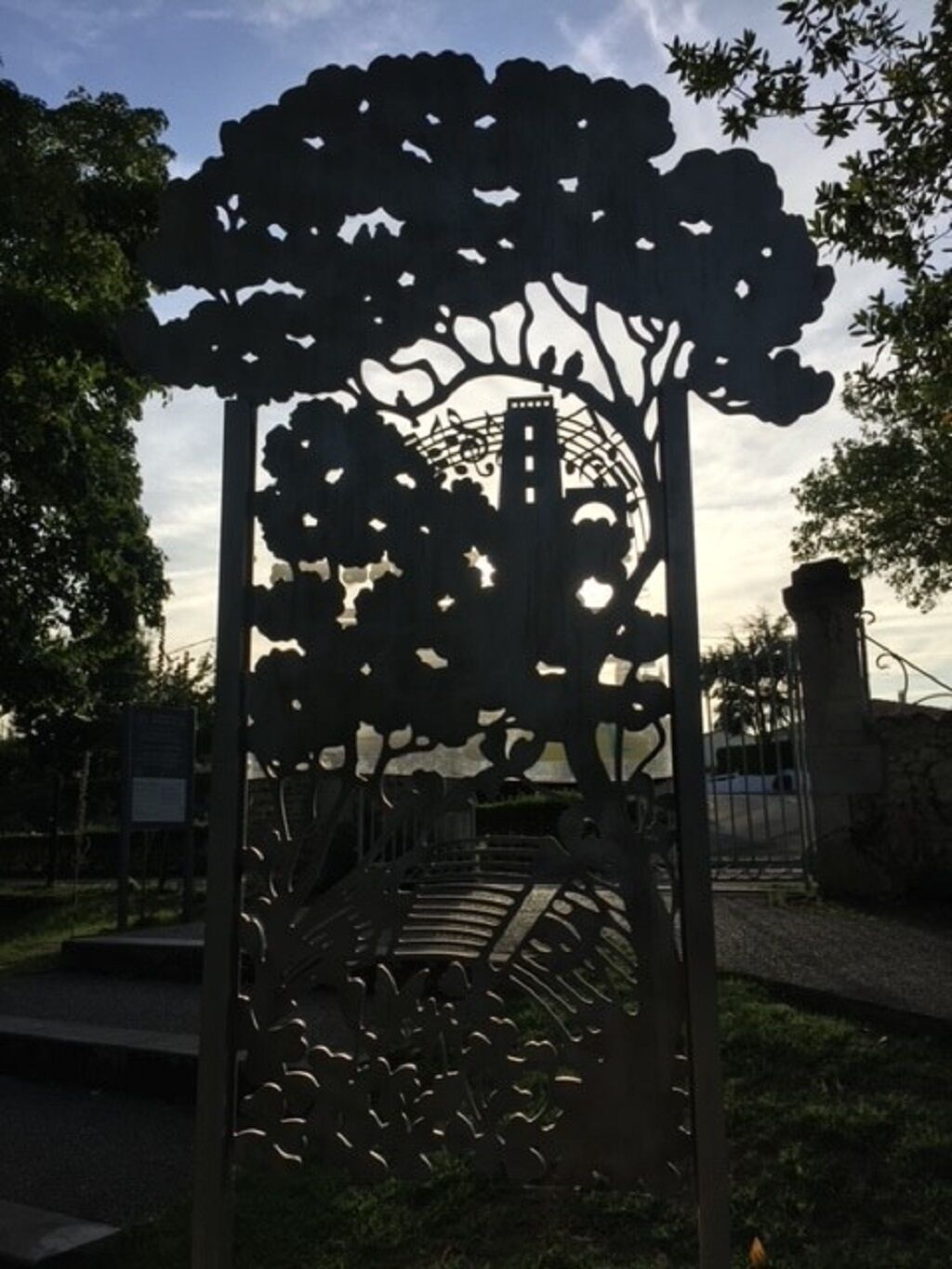 Une porte en métal ornée d’un motif découpé représentant un arbre et un pont, le tout se détachant sur un fond de feuillage et d’un ciel nuageux.