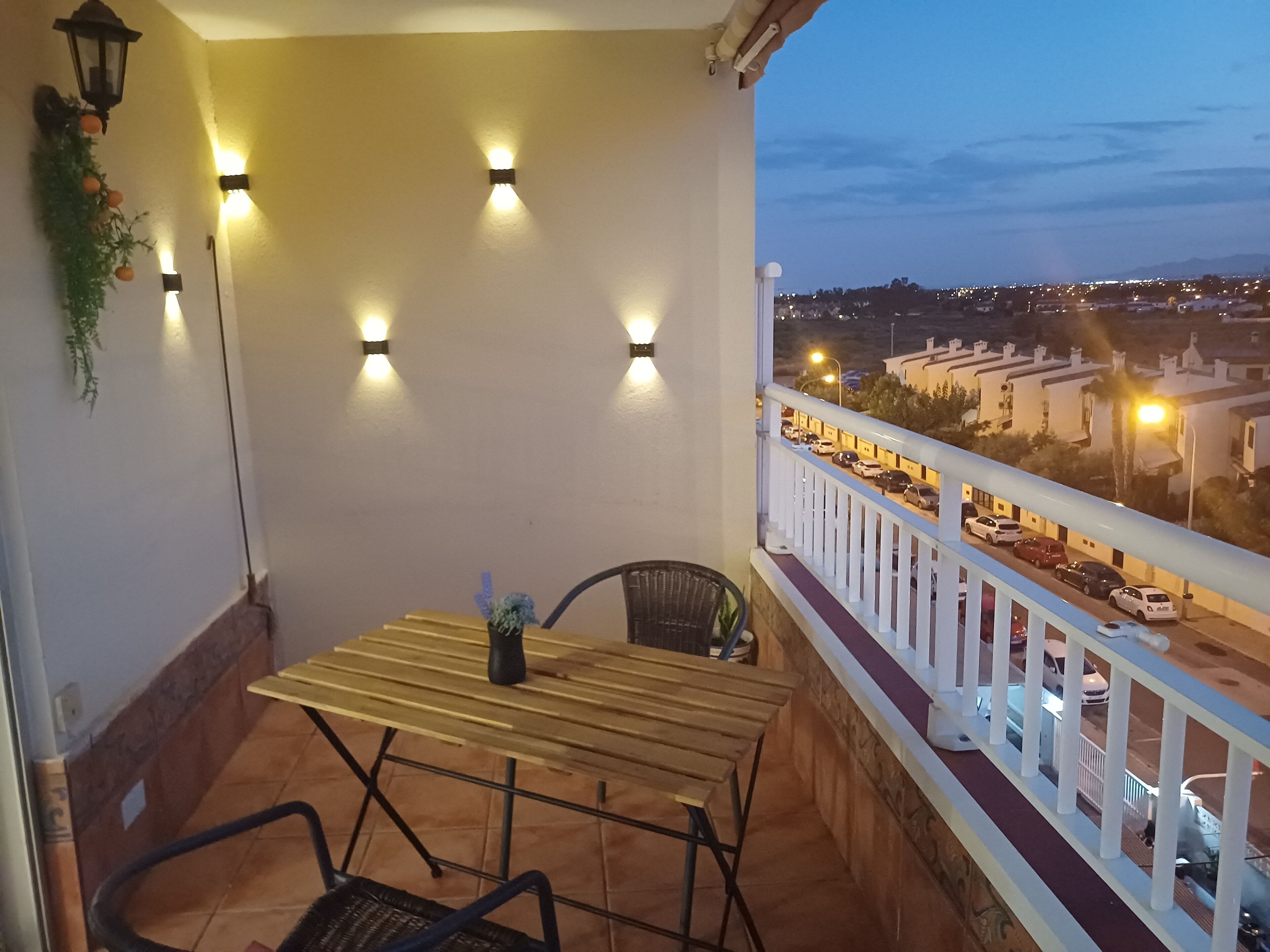 Un balcon avec une table et des chaises en bois, une plante en pot et des lampes fixées au mur.