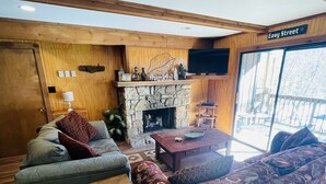 A cozy living room with a stone fireplace, wooden furniture, and a large window offering a view of the outside. - Creekside Condo Walking Distance to Slopes! (Sugar Mountain)