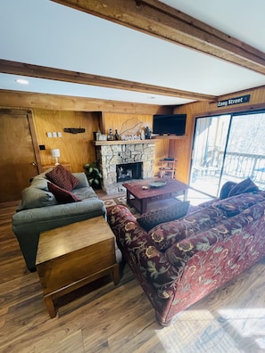 A cozy living room with a stone fireplace, wooden furniture, and a large window offering a view of the outside.