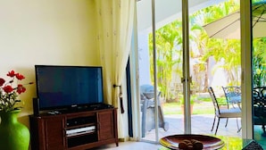 Una sala de estar con un televisor sobre un mueble de madera, una mesa de centro de vidrio con un frutero y un jarrón verde con flores rojas.