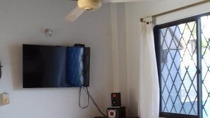 A room with a wooden ceiling, a ceiling fan, a flat-screen TV mounted on the wall, a window with a metal grid, and a table with white chairs and a fruit bowl.