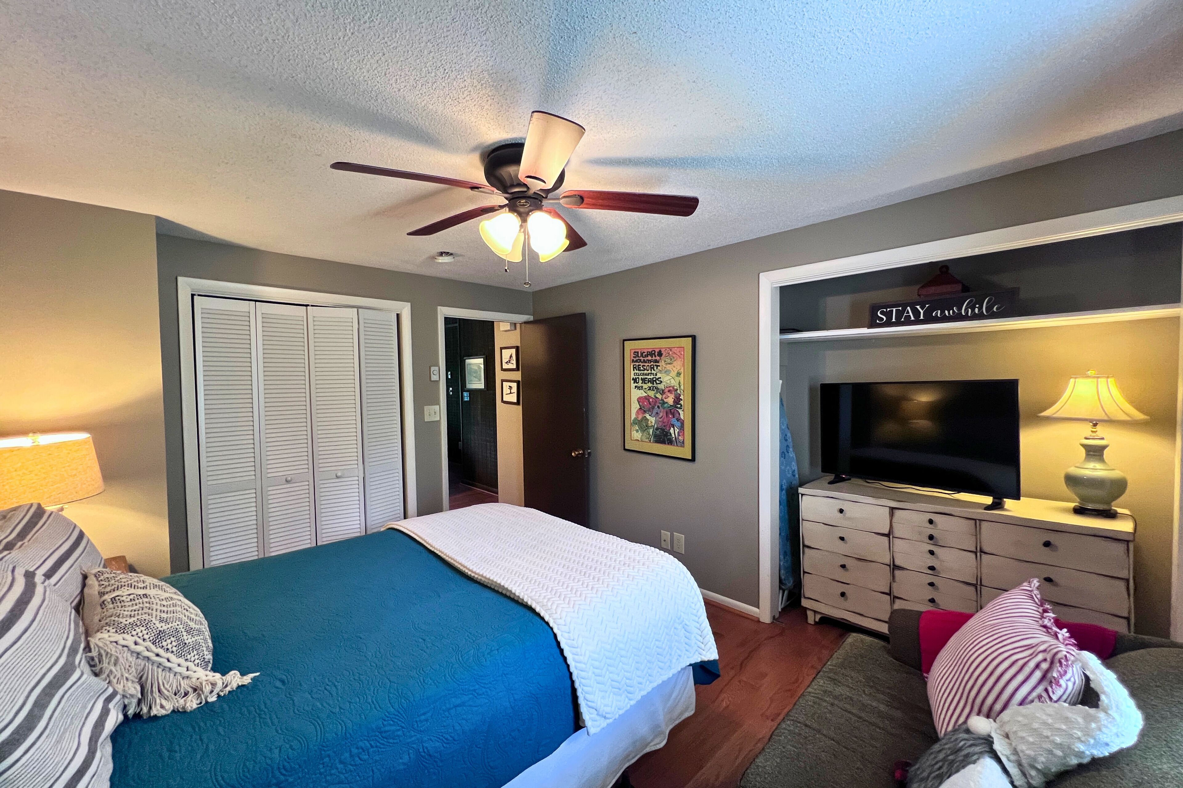 A bedroom with a bed, a ceiling fan, a television, a dresser, and a sign that says "STAY awhile".