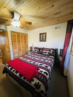 Une chambre avec un plafond en bois, un lit recouvert d’une couvre-lit à motifs, un ventilateur de plafond, une lampe fixée au mur et une porte dotée d’un volet en bois.