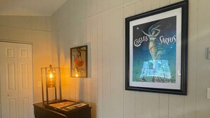 Una habitación con pared blanca con paneles, un cartel enmarcado, un escritorio con lámpara y una puerta.