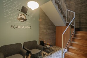 Ein modernes Interieur mit einer Treppe, zwei grauen Sesseln, einem kleinen Tisch mit einer Tasche sowie einer Wand mit Text und einer Schnurrbartgrafik.