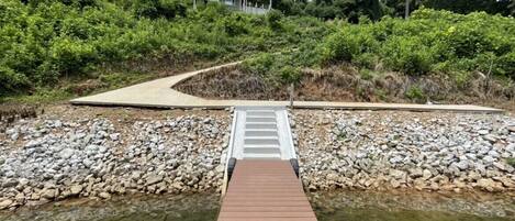 A wooden bridge over a river with a pathway leading to a building surrounded by greenery.