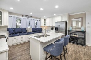 Une cuisine moderne avec des meubles blancs, un îlot central doté de tabourets de bar, un réfrigérateur en acier inoxydable et une enseigne indiquant « MAISON DE PLAGE ».