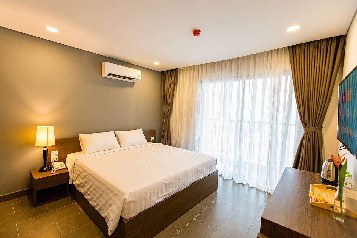A hotel room with a large bed, bedside table with a lamp, an air conditioning unit, and a window with curtains.