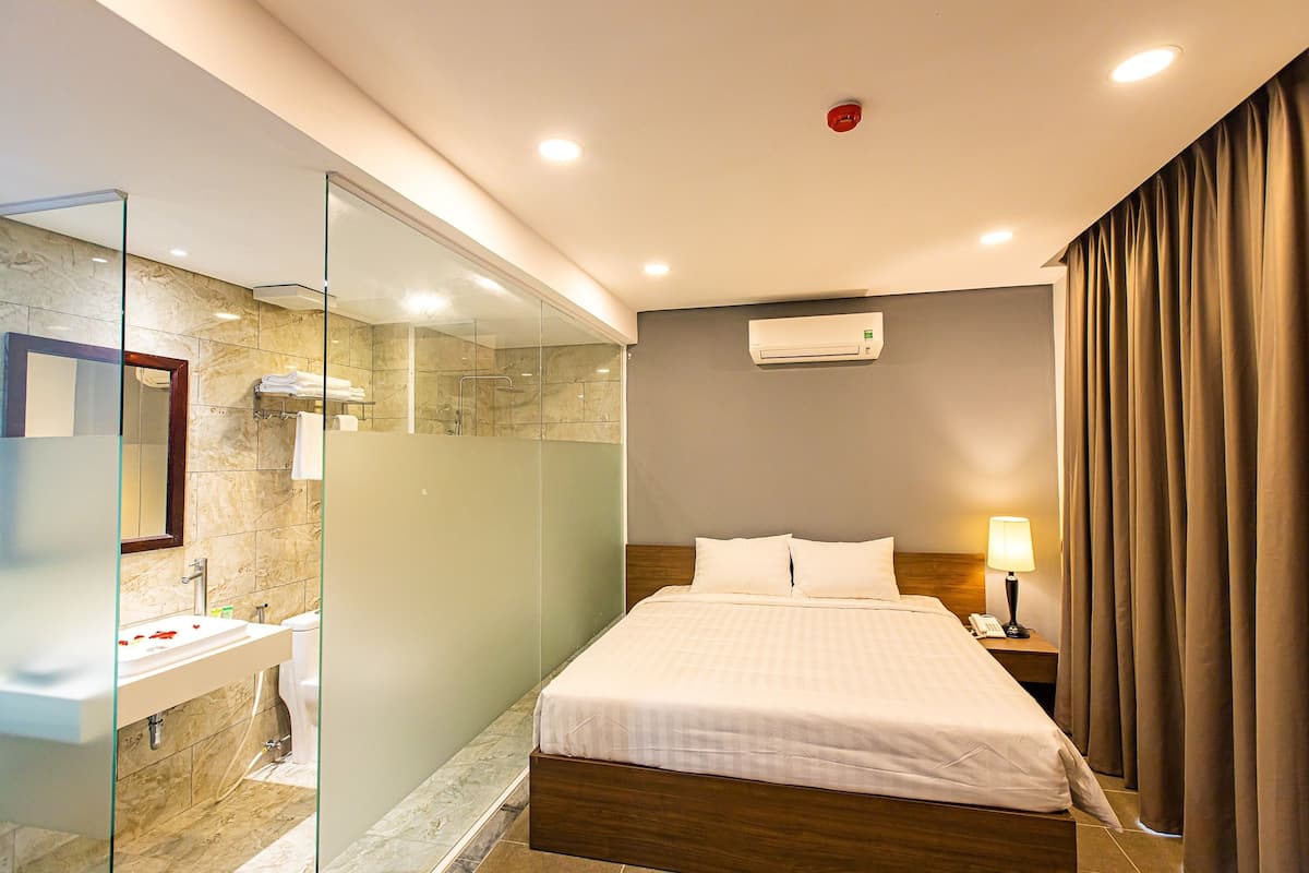 A modern hotel room with a large bed, a bathroom with a glass shower enclosure, and a mounted air conditioner.