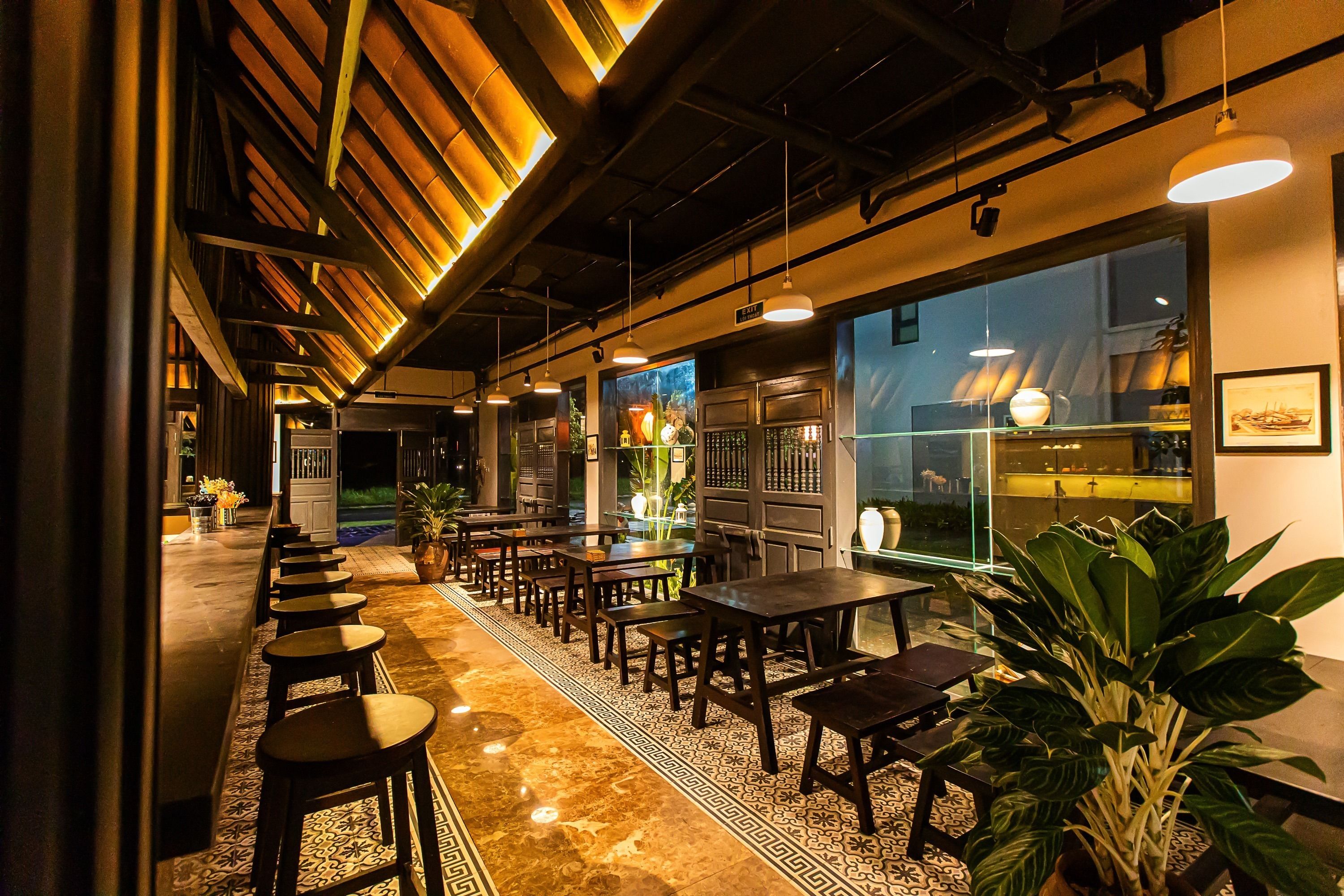 A restaurant with a long counter, multiple tables, and chairs. There are potted plants and a large window with a view of the outside.