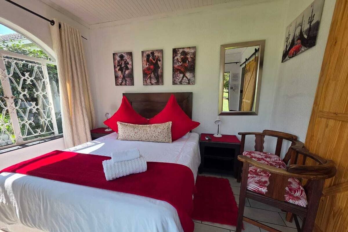 A bedroom with a large bed, red pillows, a wooden nightstand, a chair, and artwork on the wall.