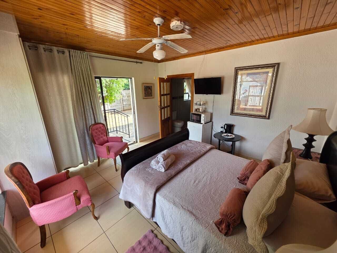 A bedroom with a bed, two chairs, a ceiling fan, a TV, and a window with curtains.