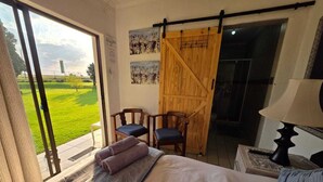 A bedroom with a bed, a chair, a wooden door, and a view of a green field. - Aloe Tree (Krugersdorp)