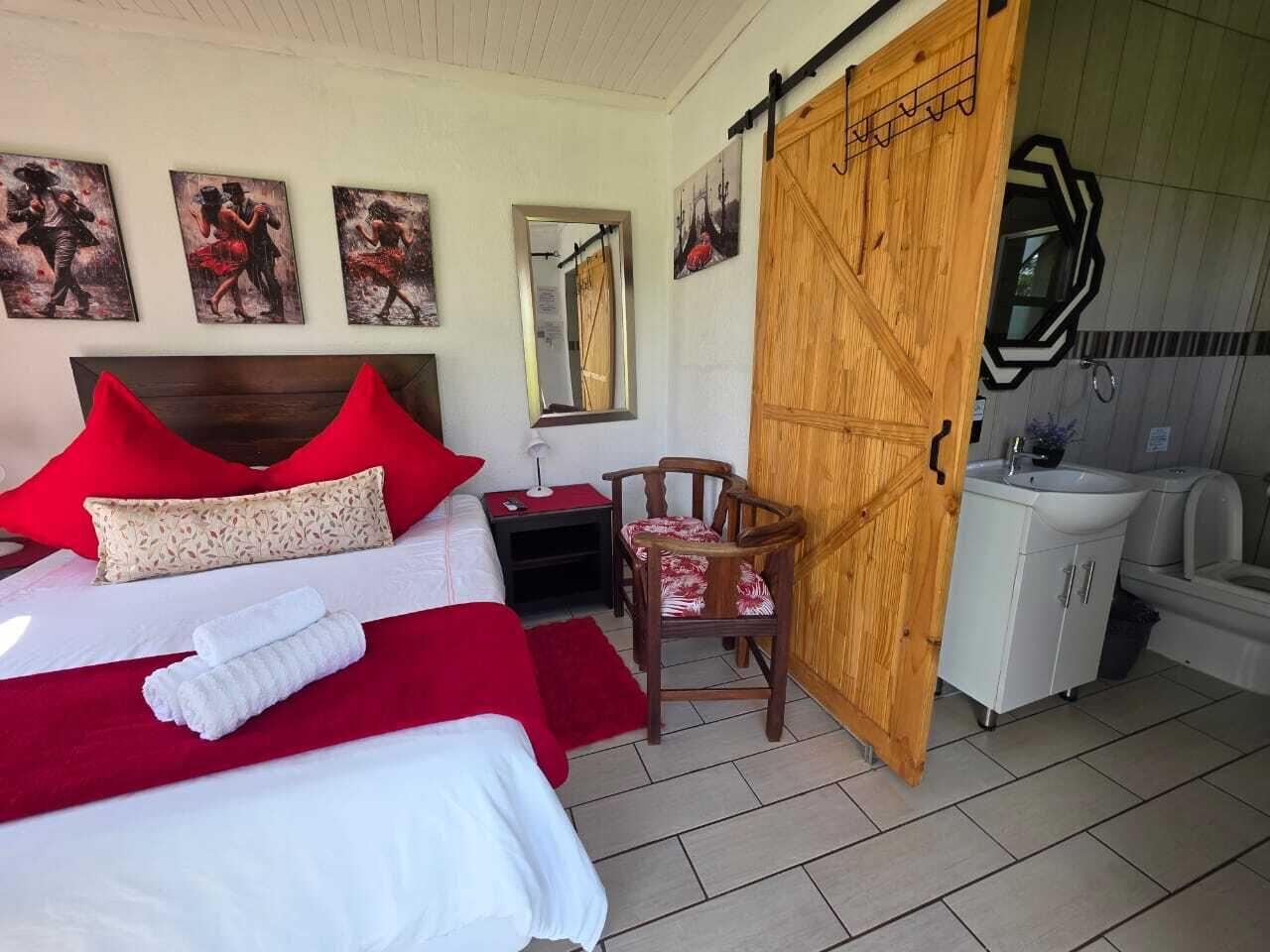 A hotel room with a bed, a wooden wardrobe, a small table with a chair, a mirror, and a bathroom with a sink and toilet.
